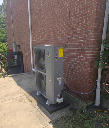Outdoor HVAC unit beside a brick building on a concrete pad with pipes and a hose nearby