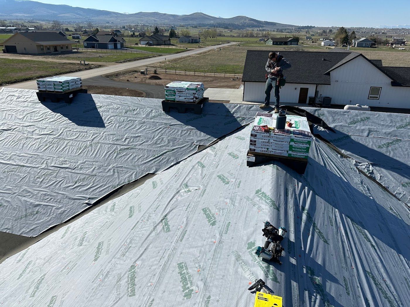 A roofer walks on a house roof covered in gray underlayment, with bundles of roofing materials placed nearby.