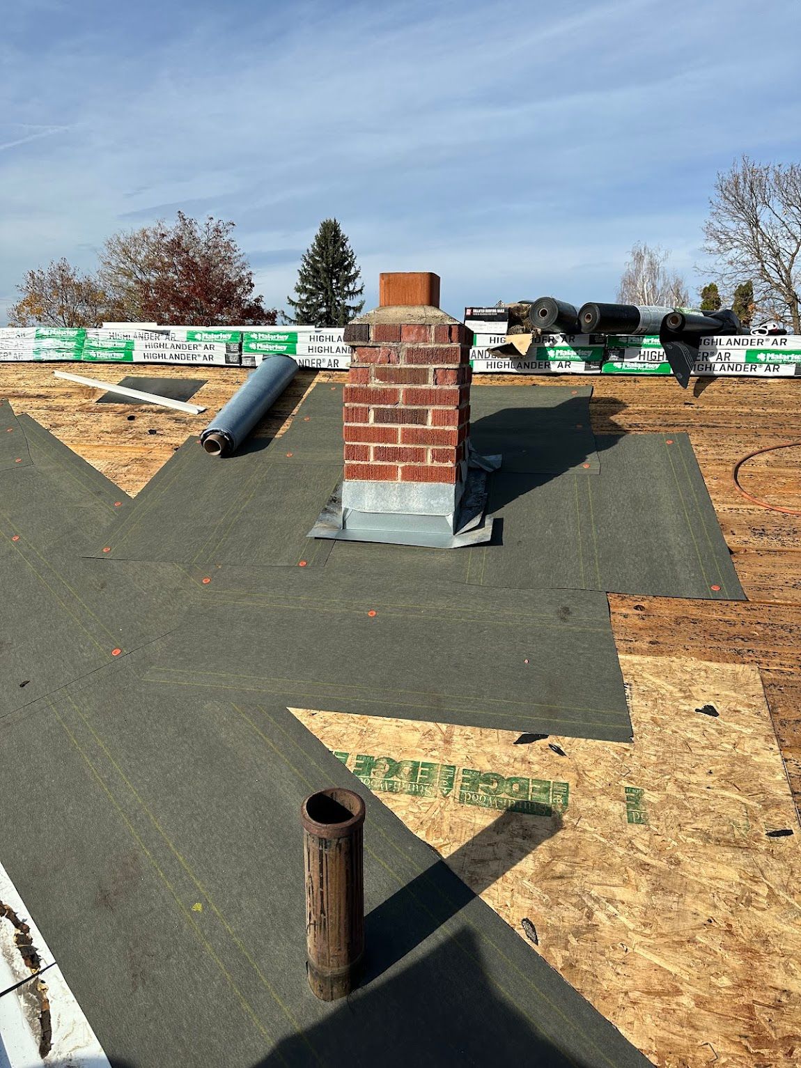 A brick chimney with metal flashing stands on a partially shingled roof under a clear blue sky.