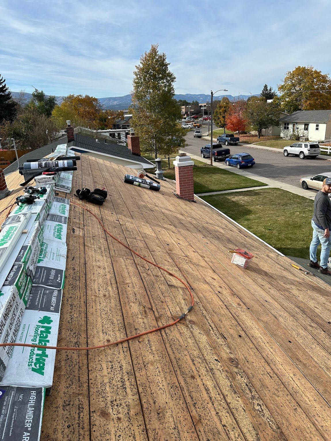 A roof under construction featuring exposed wood decking, underlayment rolls, a brick chimney, and a worker on the right.