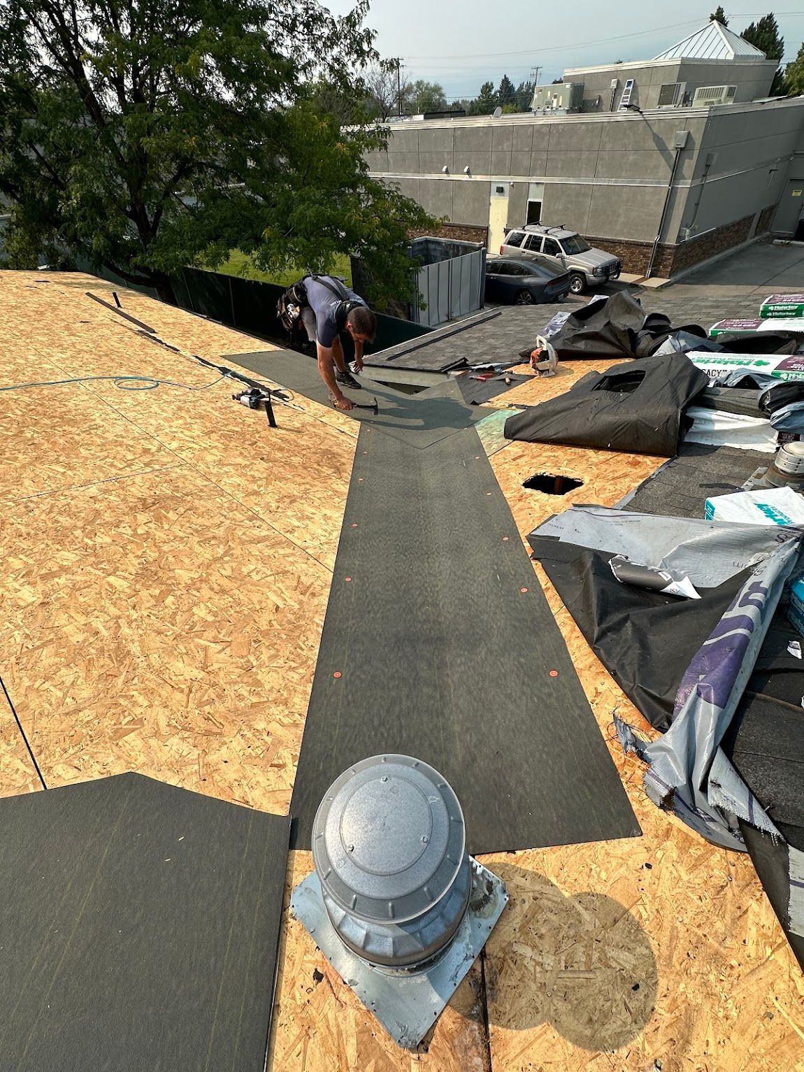 A construction worker installing roofing felt on a plywood-covered roof next to a circular roof vent.