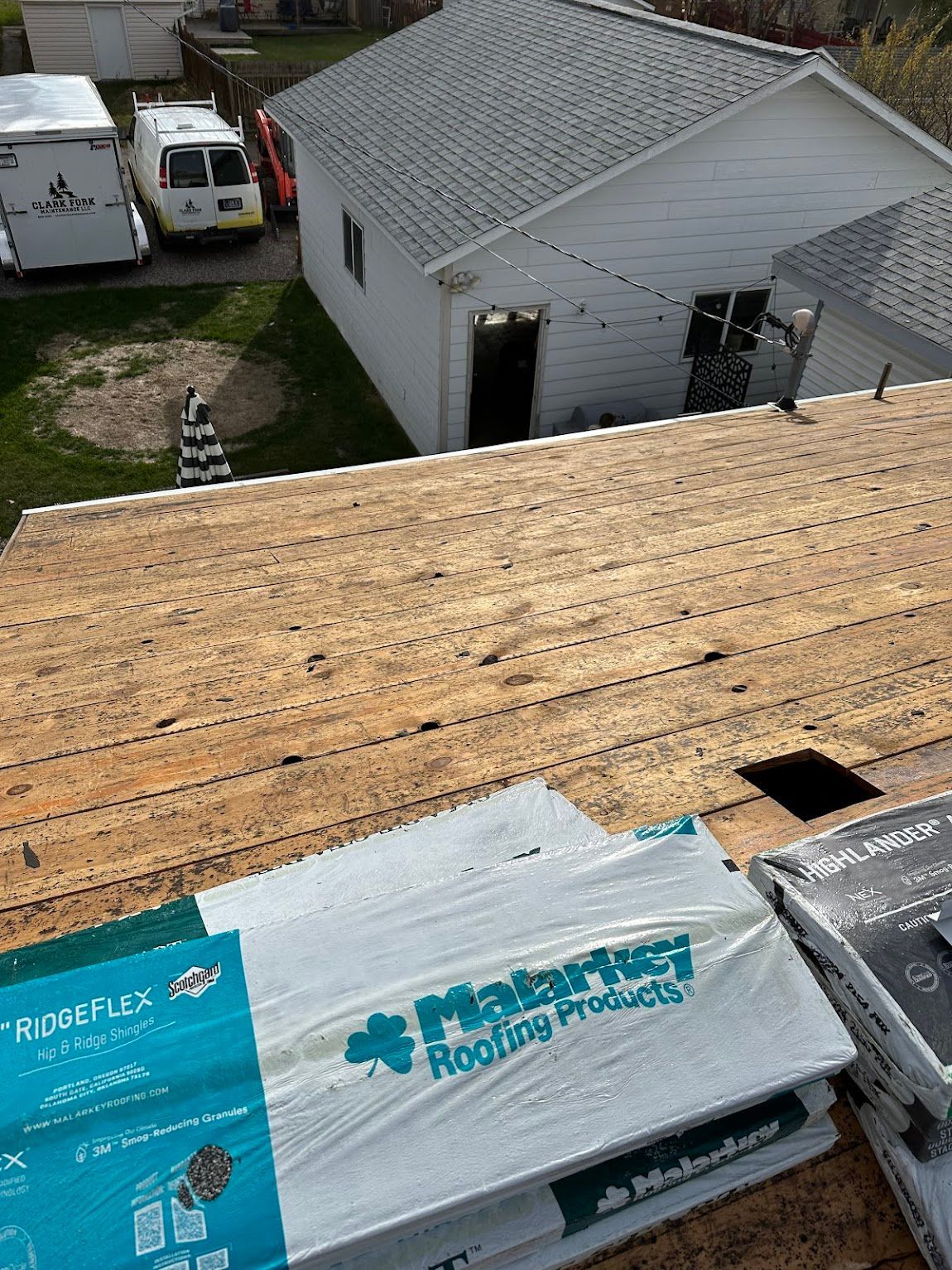 A high-angle view of a residential roof under repair, showing exposed plywood decking and stacks of roofing materials.