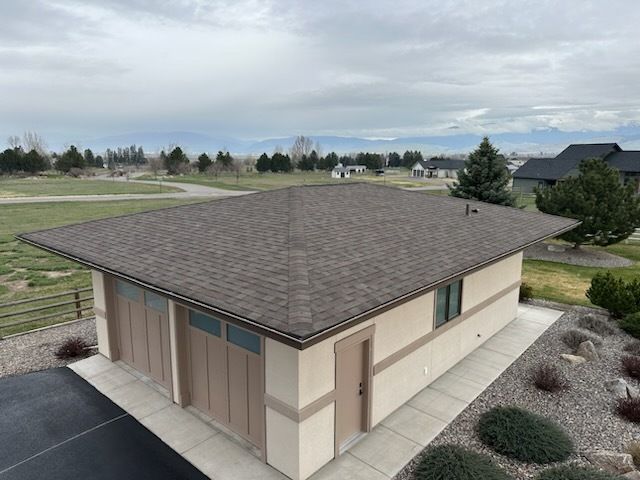 A high-angle view of a beige garage with a dark brown shingle roof, surrounded by desert landscaping and mountain views.