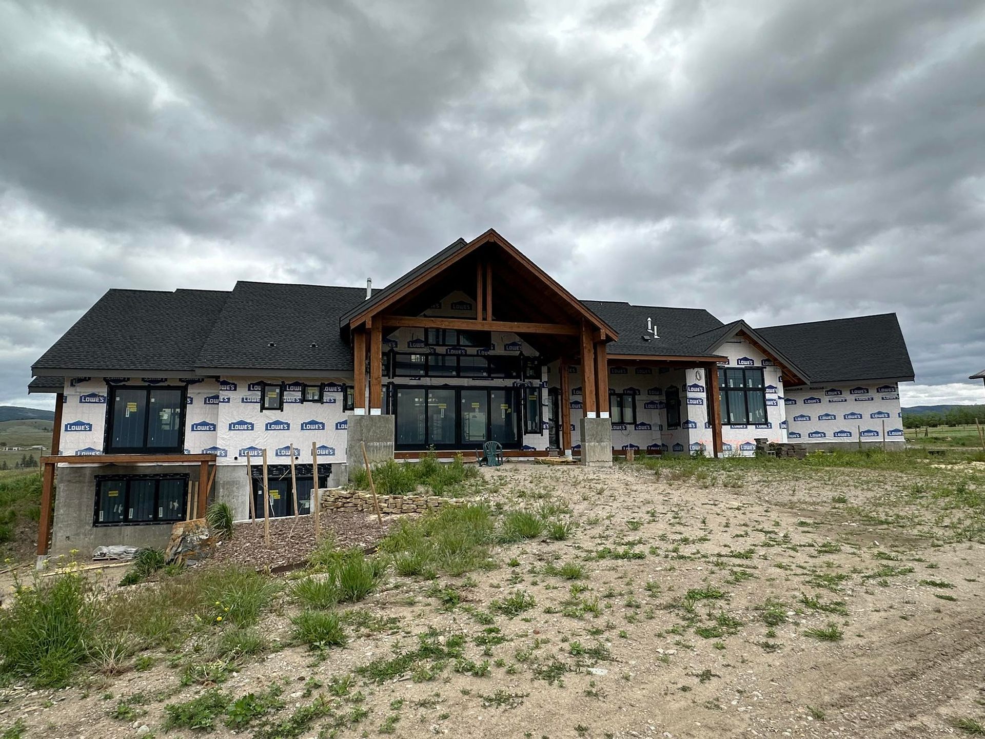 A single-story house under construction with dark shingled roofs, wood-frame entryway, and white exterior wrap.