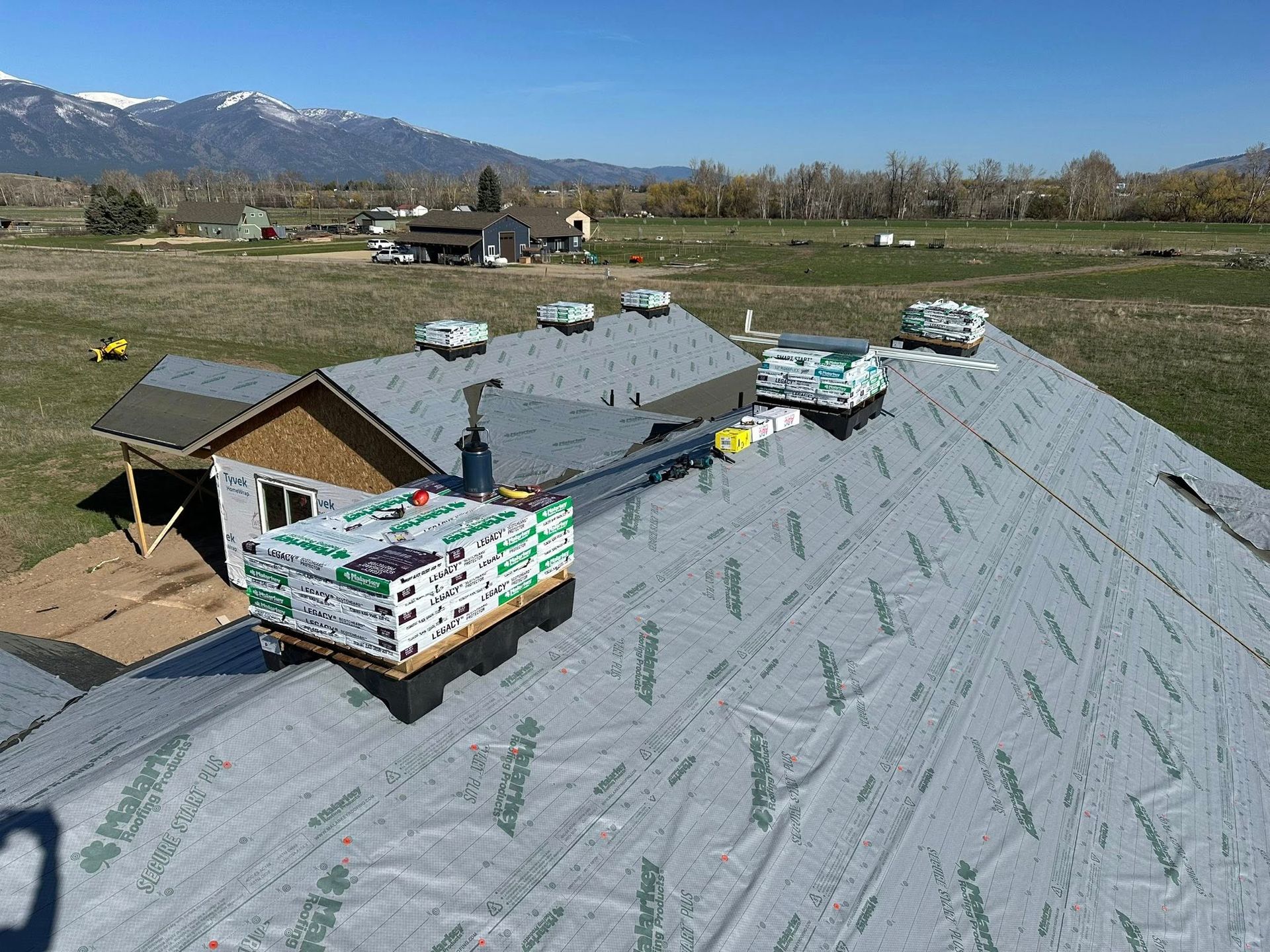 Bundles of roofing shingles sit on an underlayment-covered roof in a rural setting with mountains in the background.
