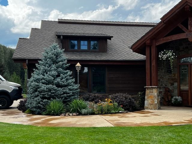 A house with dark wood siding and a stone entrance pillar, featuring a large blue spruce tree in the landscaped garden.