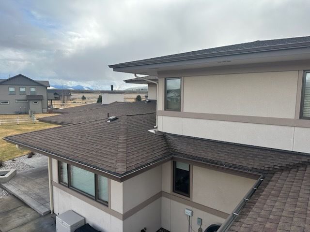 A high-angle view of a modern two-story house with beige stucco walls, dark brown shingled roofs, and mountains in the back.