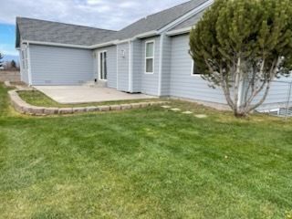 A light blue house with a gray roof features a concrete patio bordered by a low stone wall next to a grassy yard.