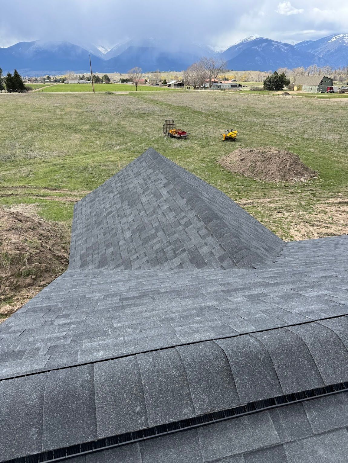 A high-angle view looking over a newly shingled gray roof toward a grassy field and distant snow-capped mountains.