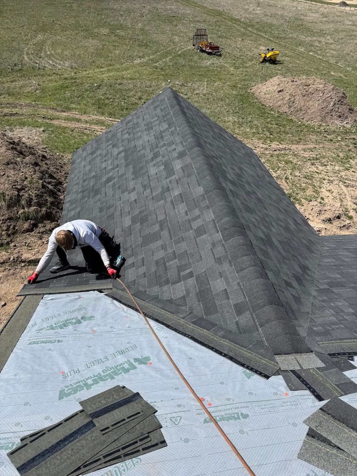 A worker in light-colored clothing installs black shingles on a roof partially covered in light gray underlayment.
