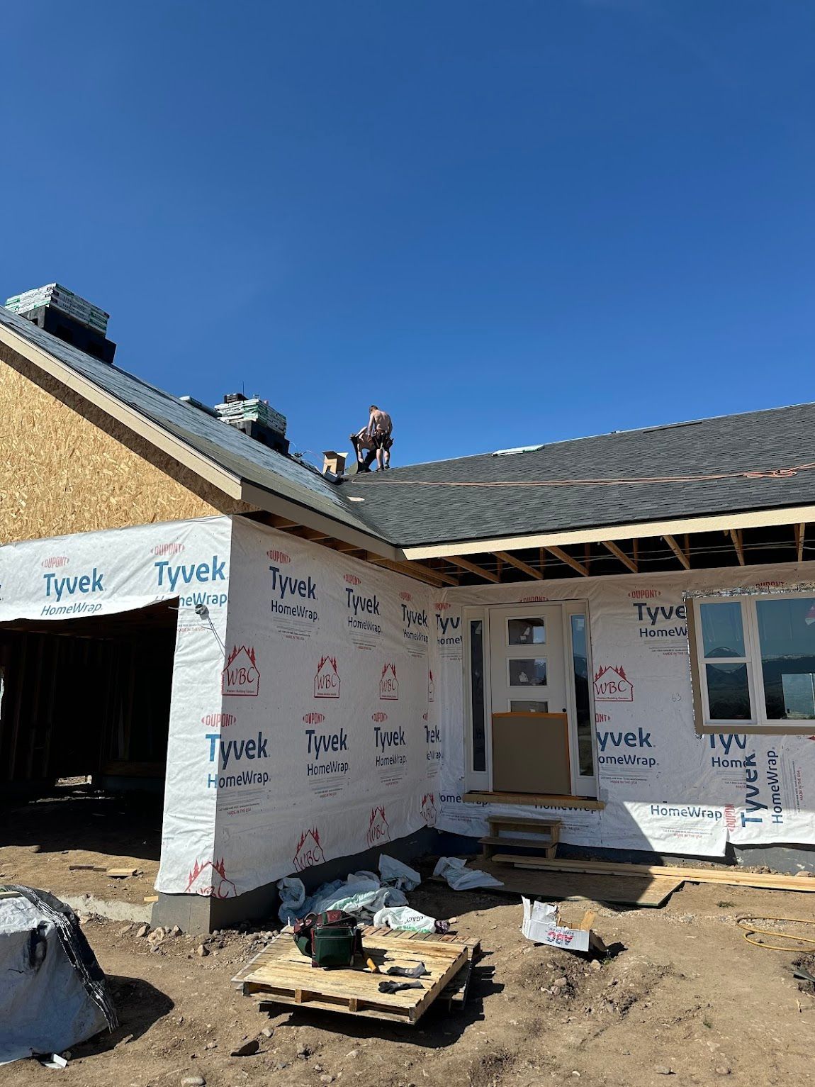 Workers install roof shingles on a house under construction wrapped in white Tyvek building paper.