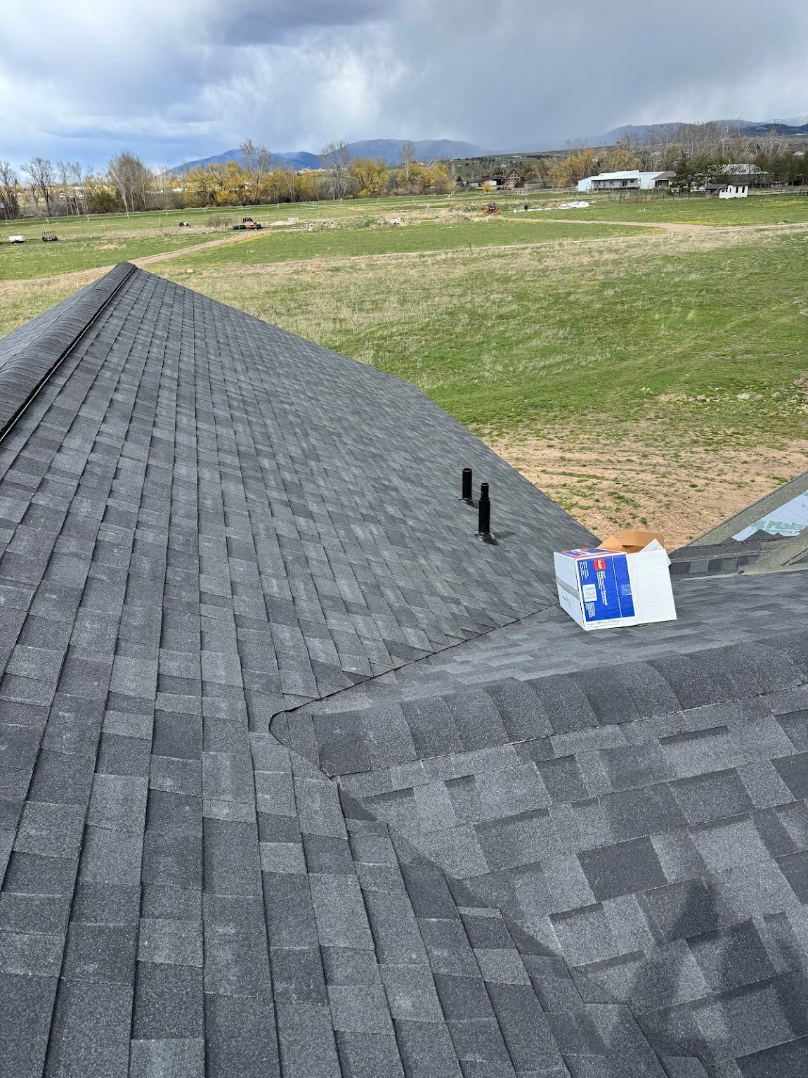 View from a residential roof overlooking a grassy field under a cloudy sky, with a box of roofing shingles nearby.