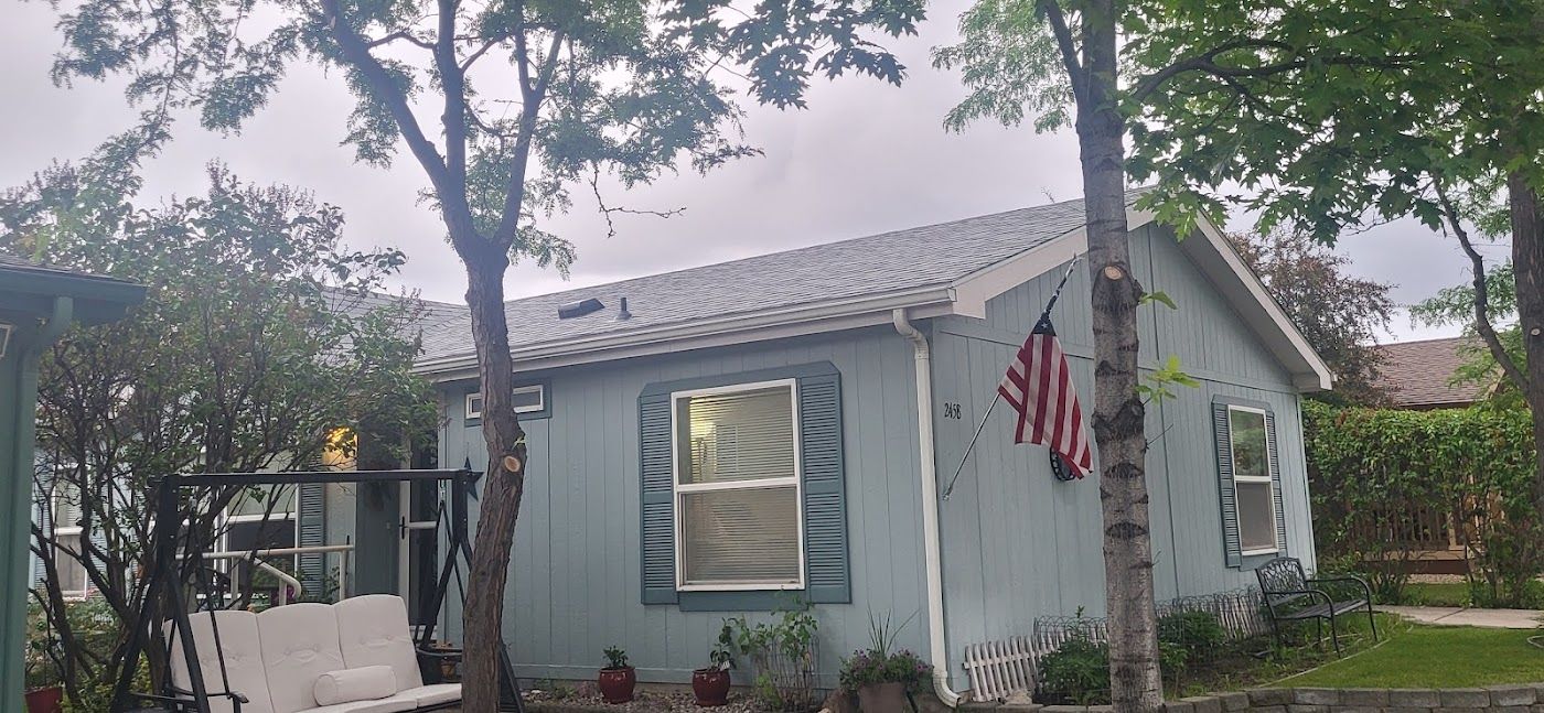 A light blue single-story home with a white-trimmed roof, blue shutters, and an American flag hanging by the window.