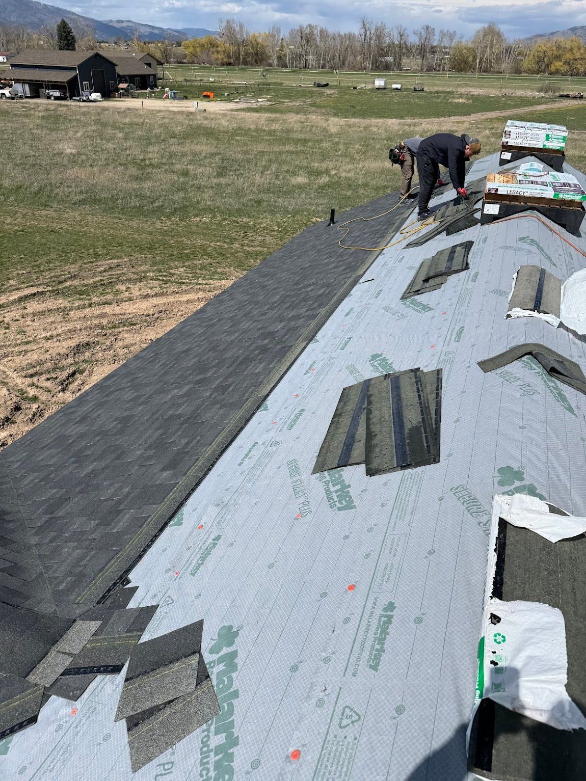 A construction worker installs black shingles onto the underlayment of a residential roof in a rural, sunny setting.