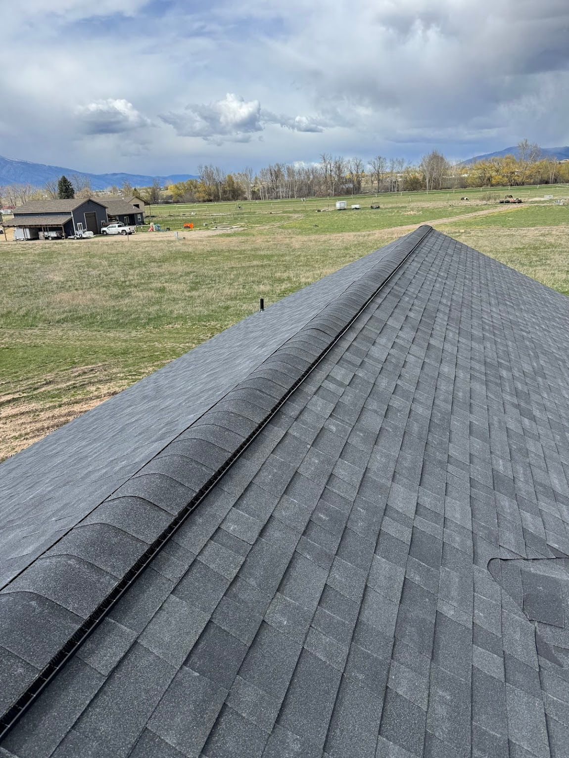 A high-angle view looking down the gray shingled ridge of a roof toward a rural field and distant mountains.