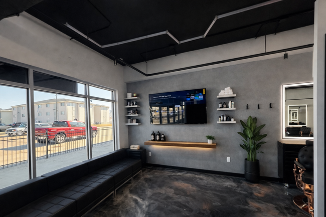 Modern salon waiting area with a black bench, gray walls, a wall-mounted TV, shelves, and a large mirror.