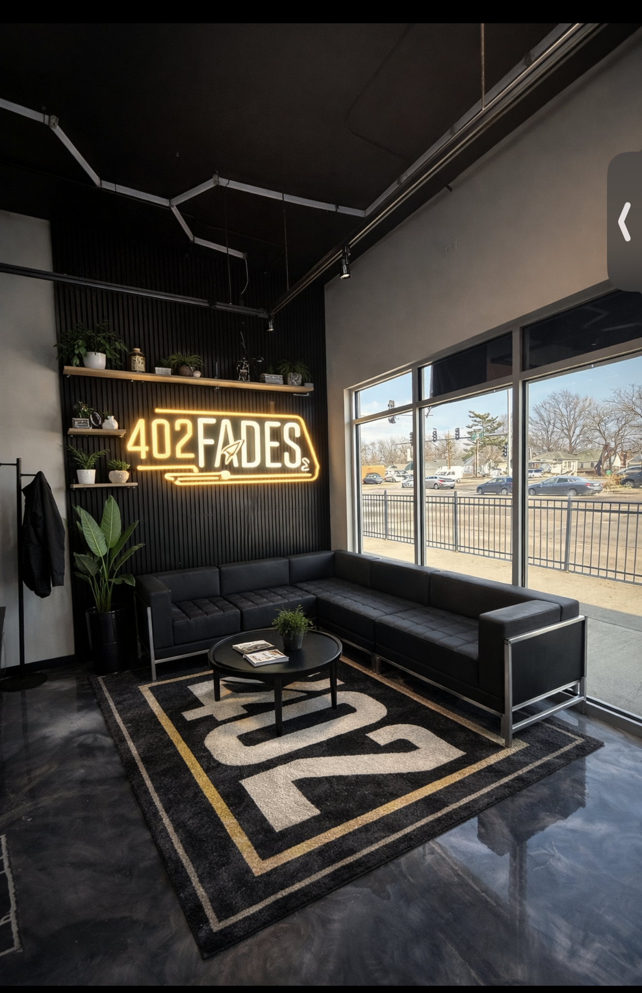 A modern barber shop waiting area with a black L-shaped sofa, a "402 FADES" neon sign, and a matching logo area rug.
