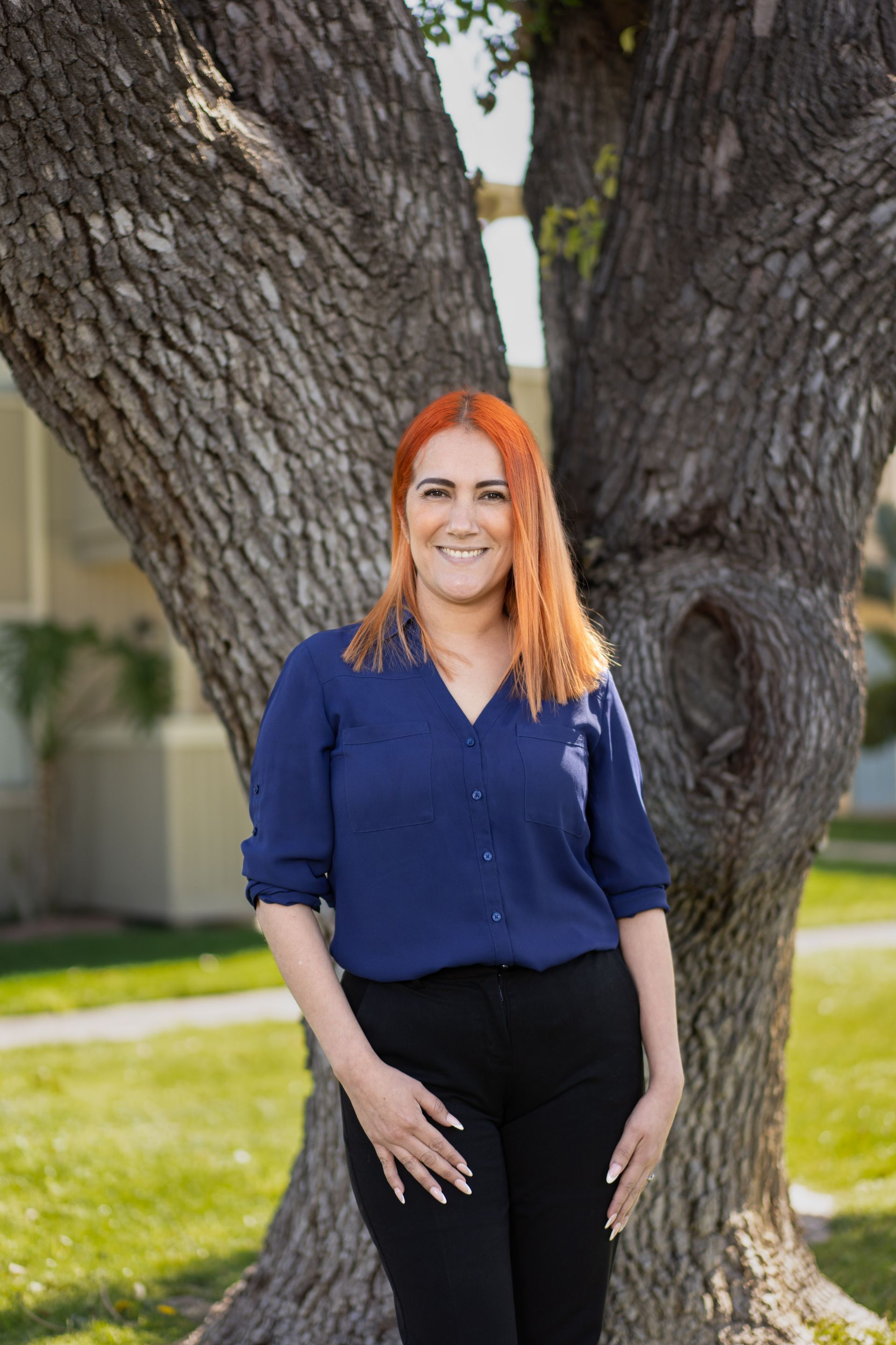 Woman with red hair smiles, leans against a tree, wearing a blue shirt and black pants outside.