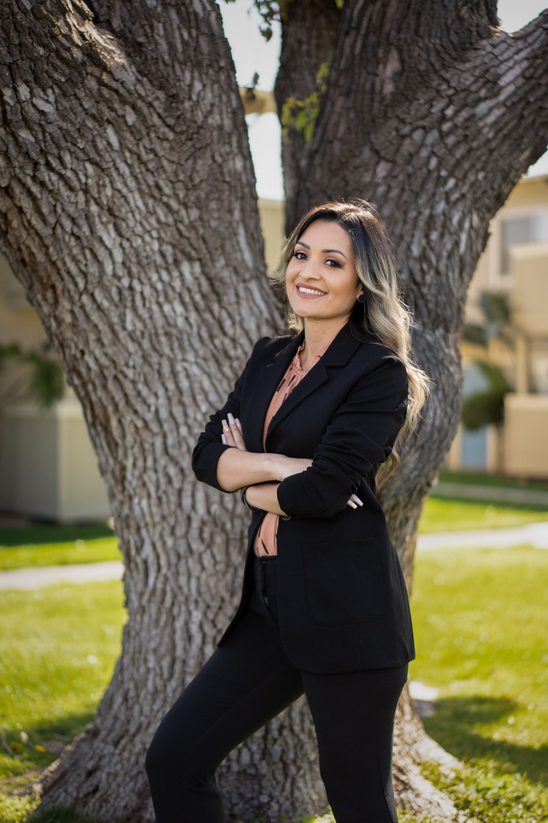 Woman in a black blazer and pants, arms crossed, smiling in front of a tree.