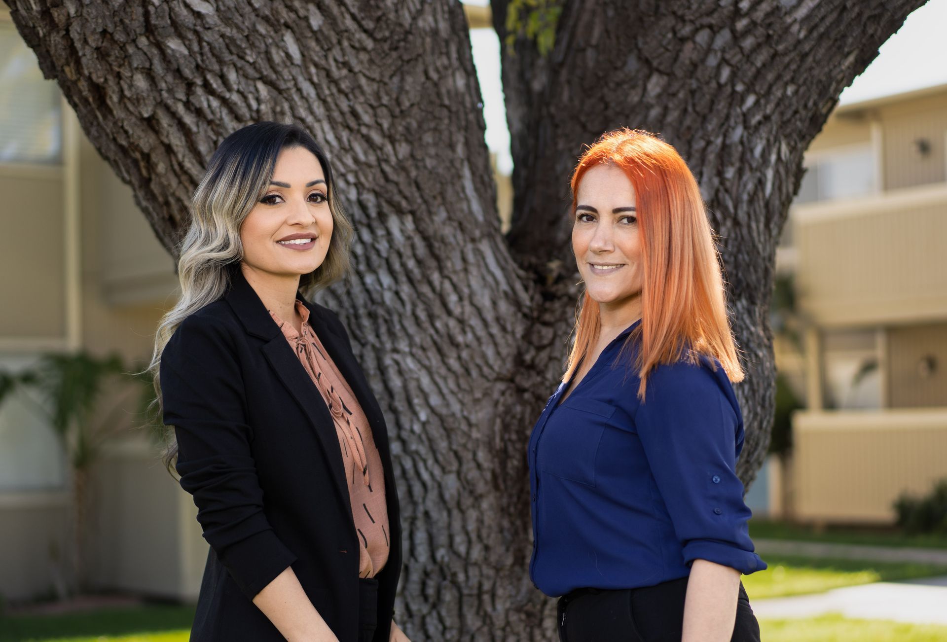Two women stand smiling near a large tree, one in a blazer, the other in a blue shirt.