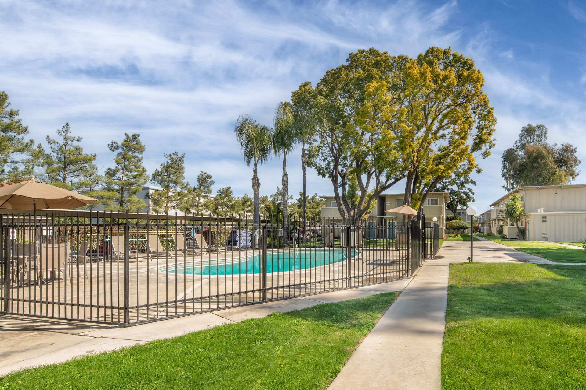 Pool area with fence, green grass, trees, and a building under a blue sky.