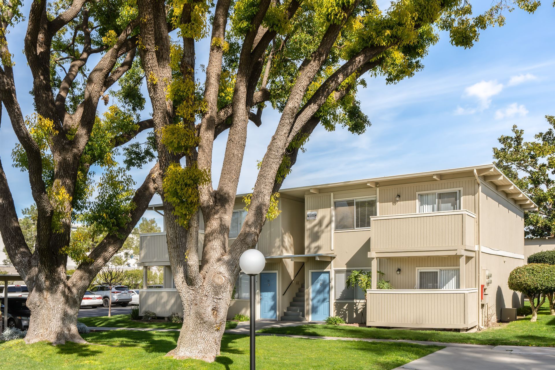 Two-story tan apartment building with blue doors, tall trees, and a grassy lawn under a blue sky.