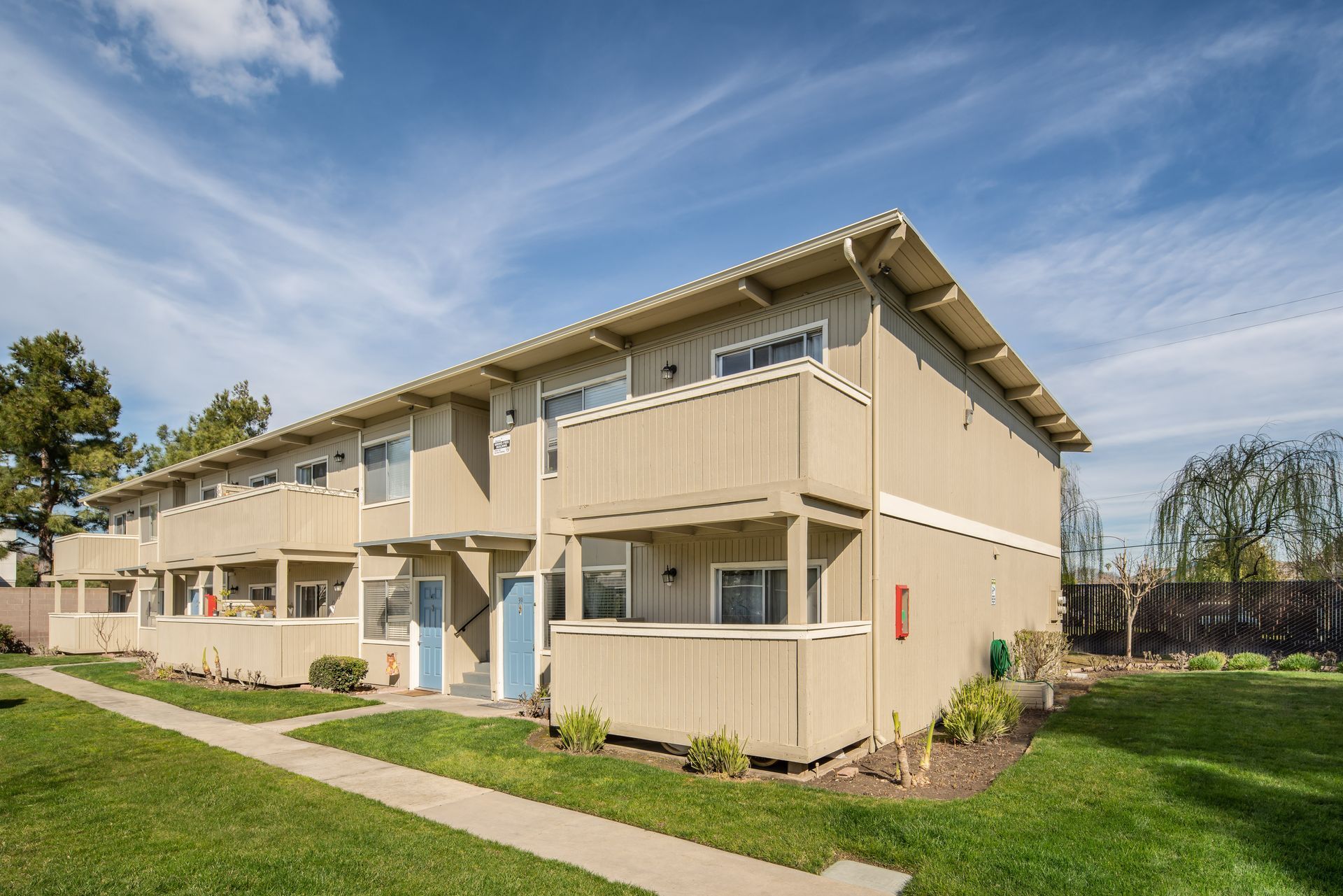 Two-story beige townhomes with balconies, blue doors, and a green lawn under a blue sky.