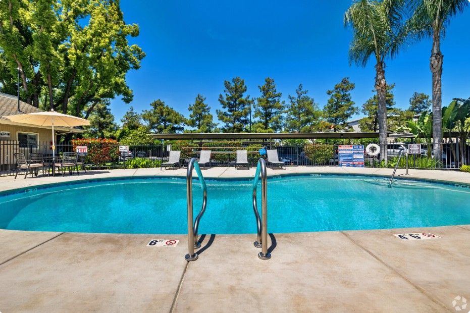 A community pool with clear blue water, surrounded by a concrete deck, chairs, and trees under a sunny, cloudless sky.