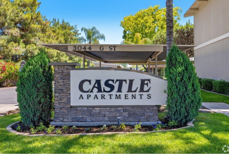 Pool area with fence, green grass, trees, and a building under a blue sky.