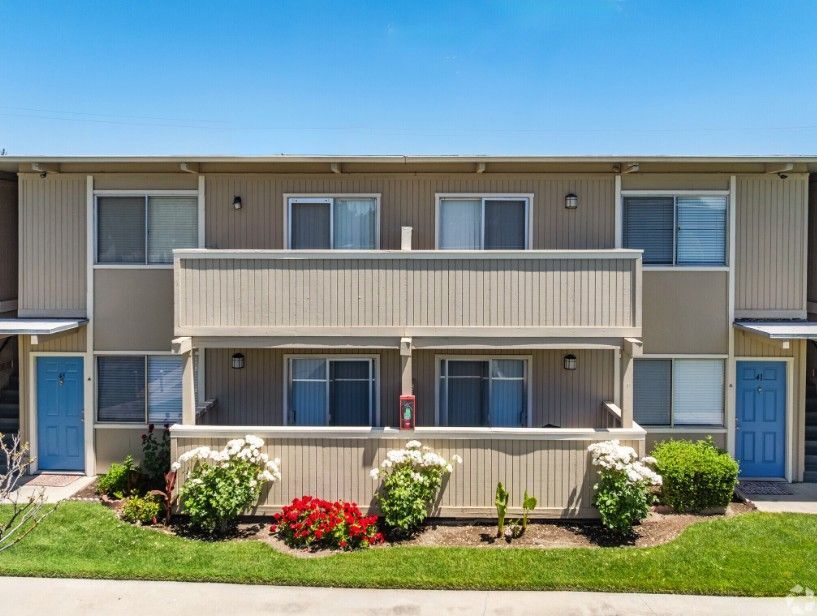 Two-story beige apartment building with blue doors, balconies, and landscaping; blue sky.