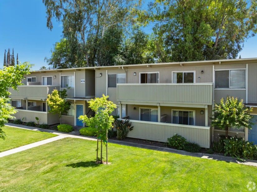 Apartment building exterior with balconies, green grass, and trees under a bright blue sky.