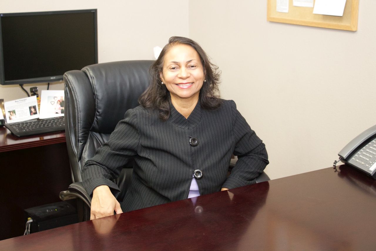 Woman in a blazer sits at a desk, smiling. Desk is in an office setting with a computer and phone.