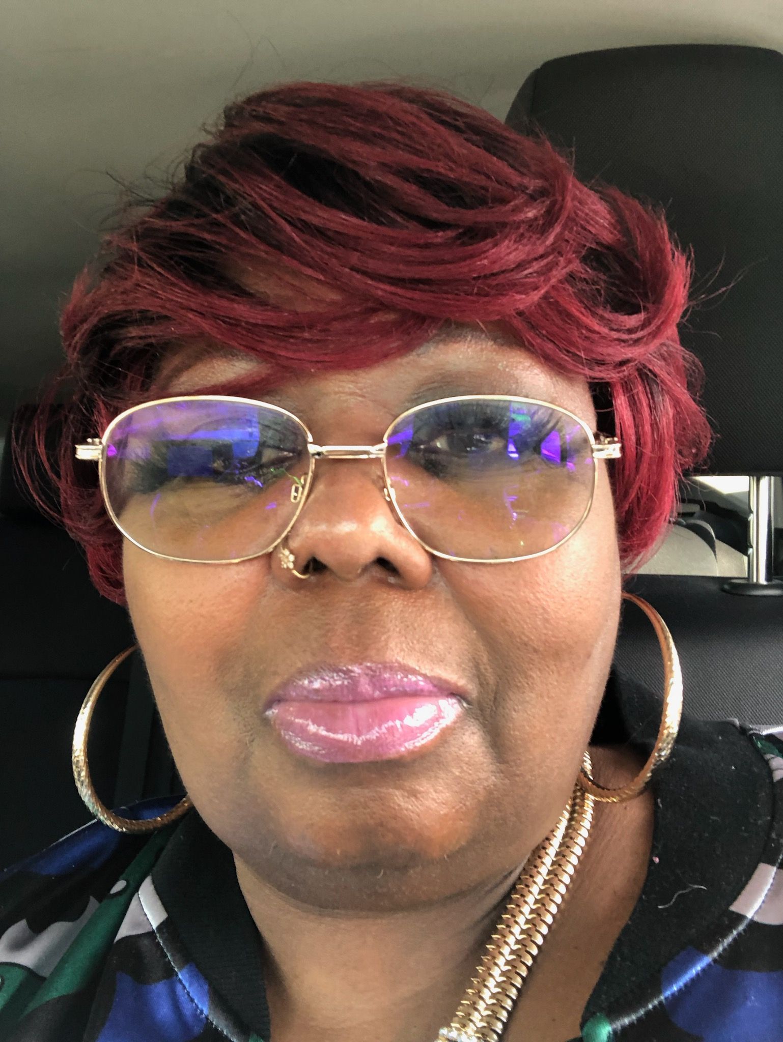 Woman with burgundy hair, gold-rimmed glasses, hoop earrings, and a nose ring, smiling, inside a car.