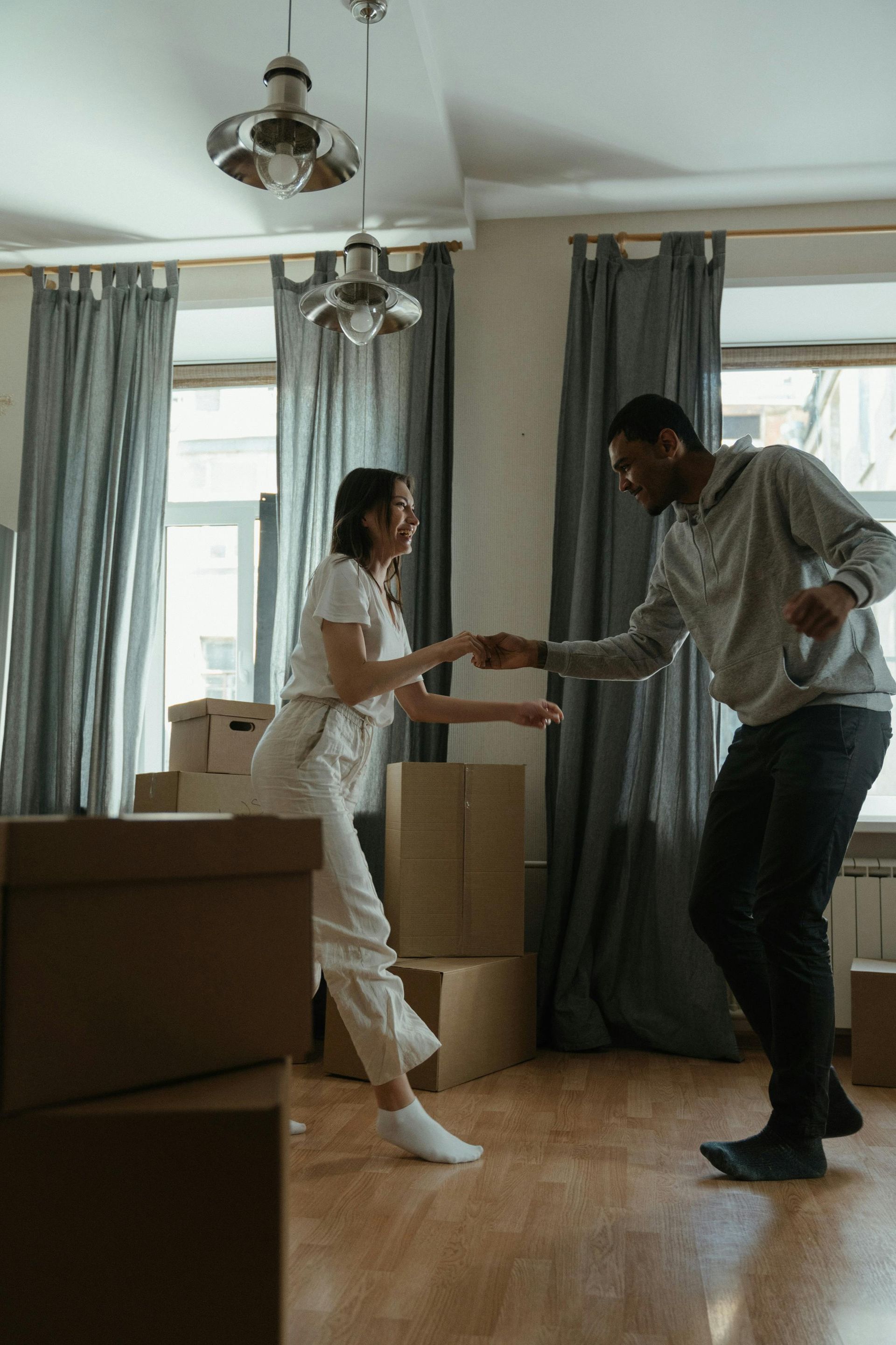 Couple dancing in a room with moving boxes. Woman in white smiling; man in gray smiling.