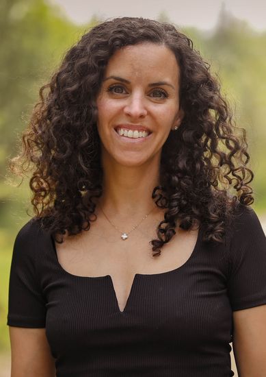 Photo of Naama smiling, wearing a black top, outdoors with blurred greenery.