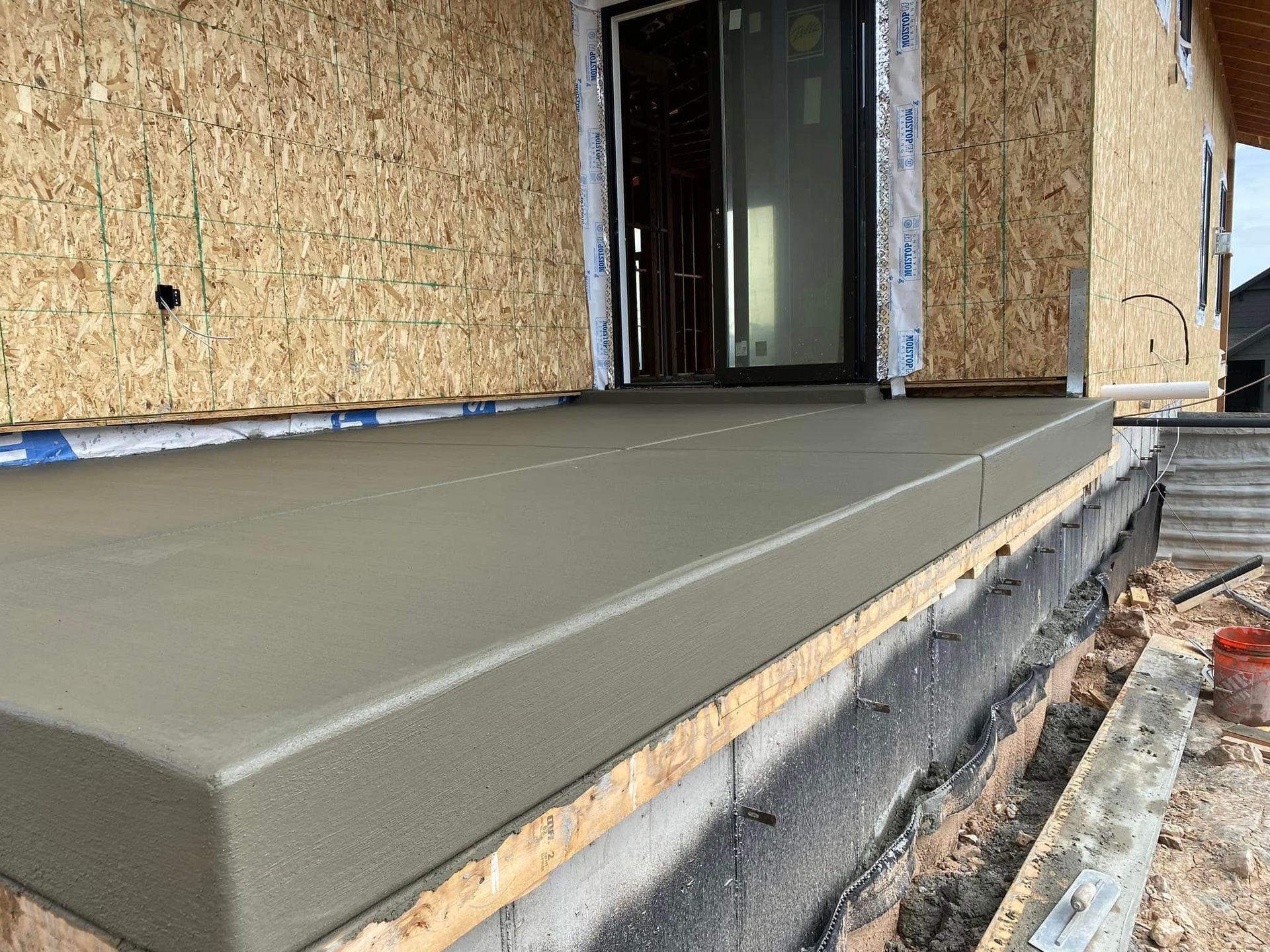 A freshly poured concrete porch sits in front of a house under construction with exposed oriented strand board walls.