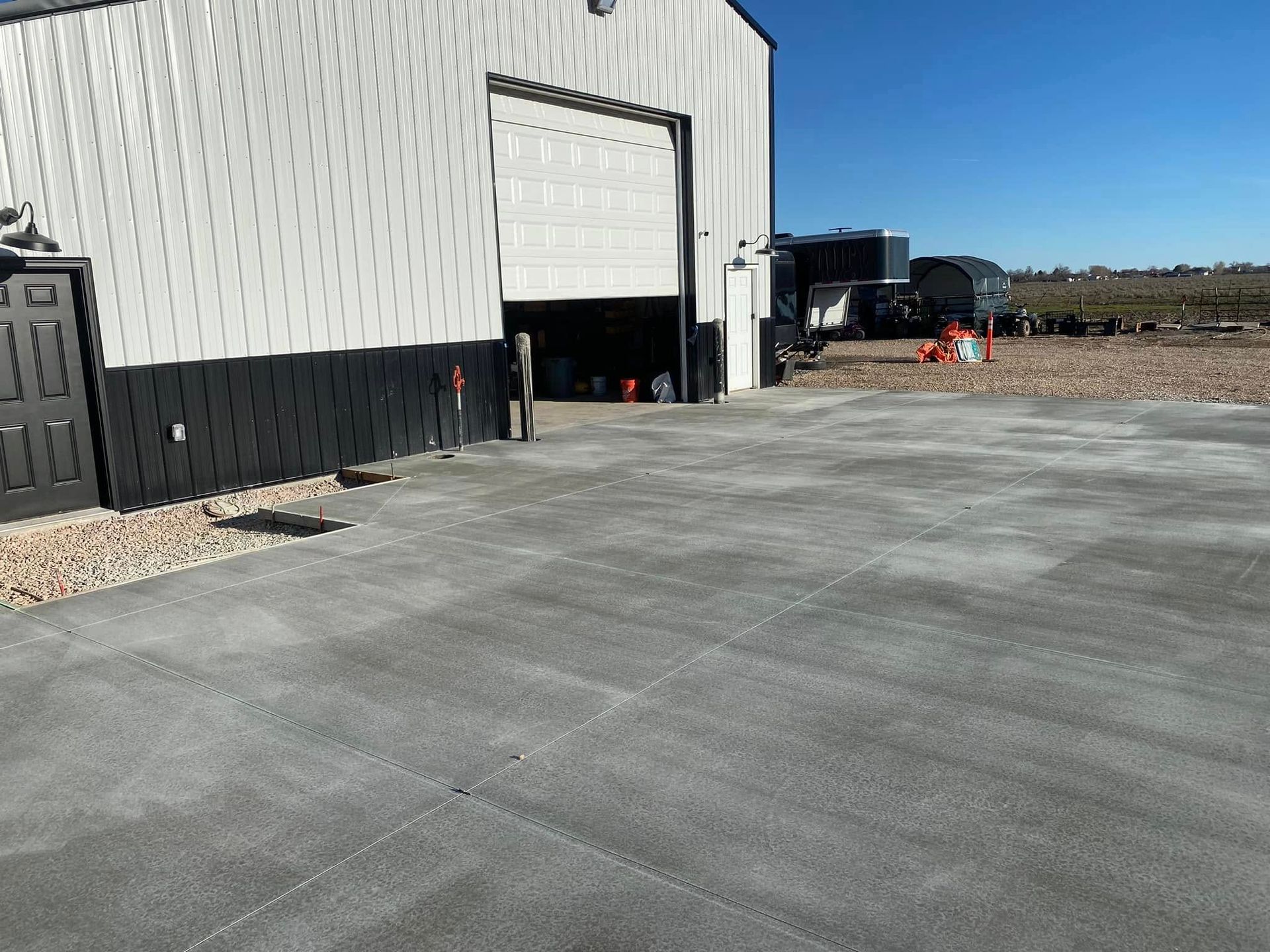 A white metal building with a large open bay door, a black entrance door, and a concrete driveway in a rural setting.