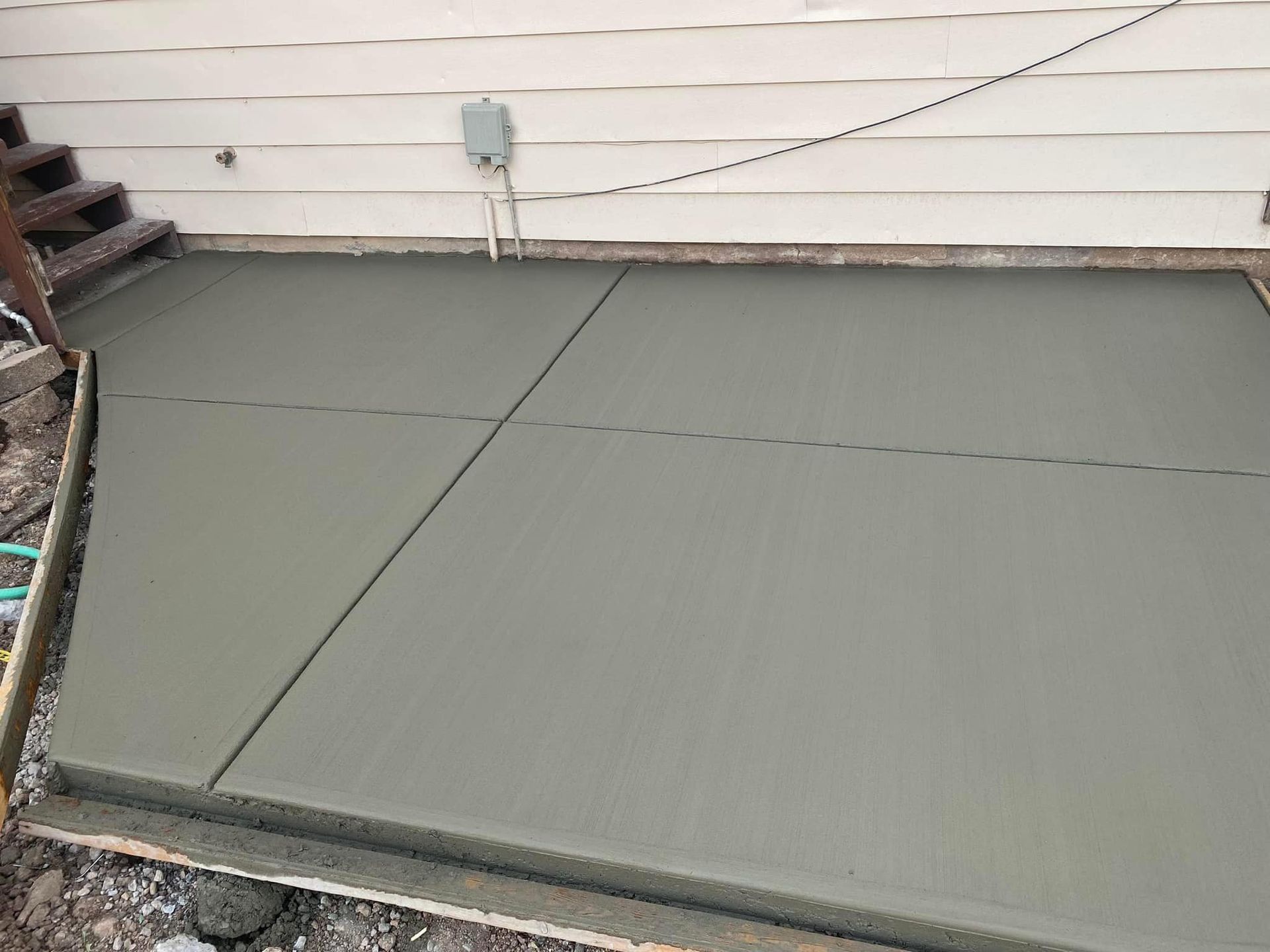 A freshly poured gray concrete patio slab next to the side of a house with wooden siding and outdoor stairs.