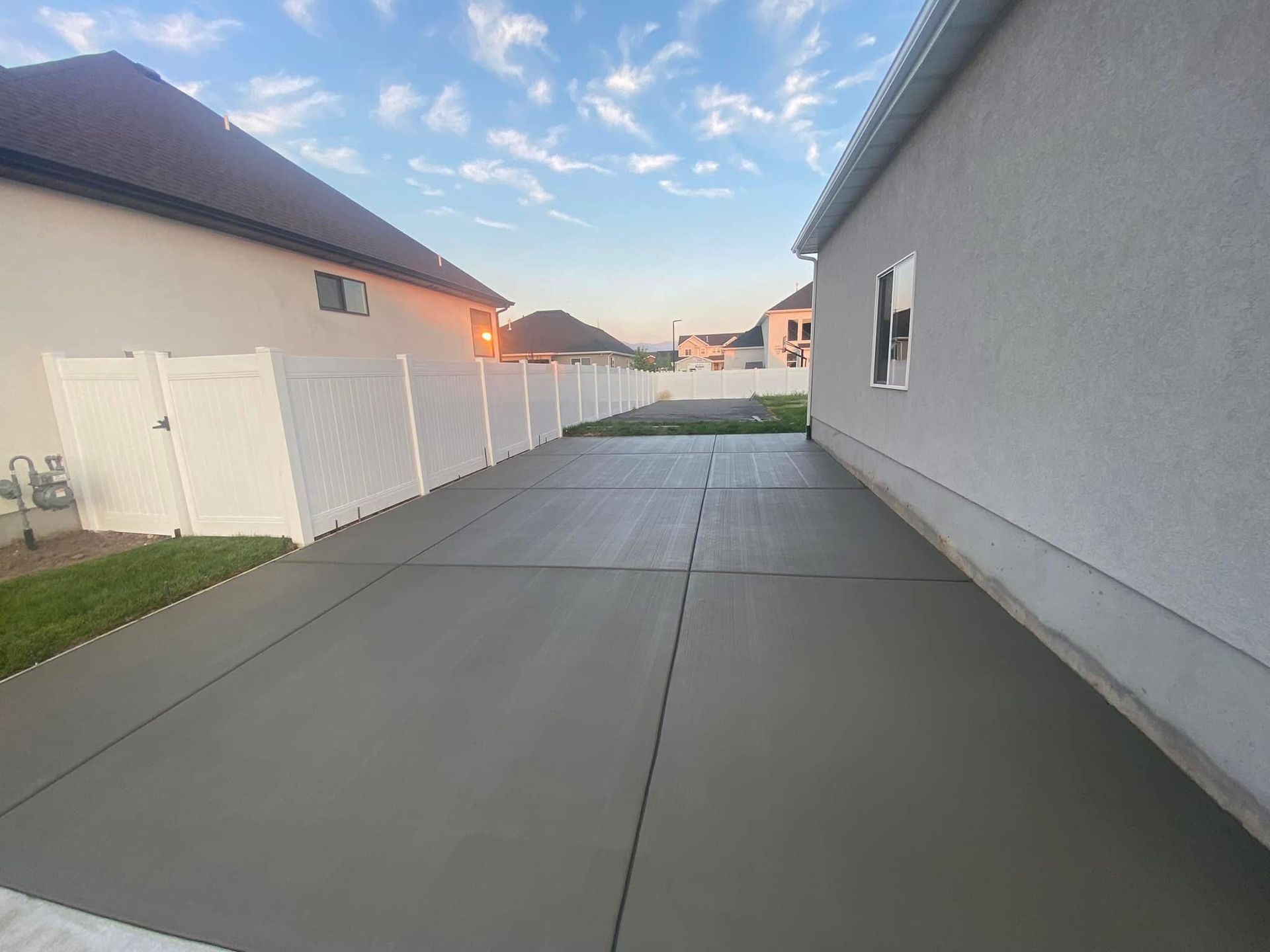 A wide concrete driveway sits between a white vinyl fence and the side of a light-colored stucco house.