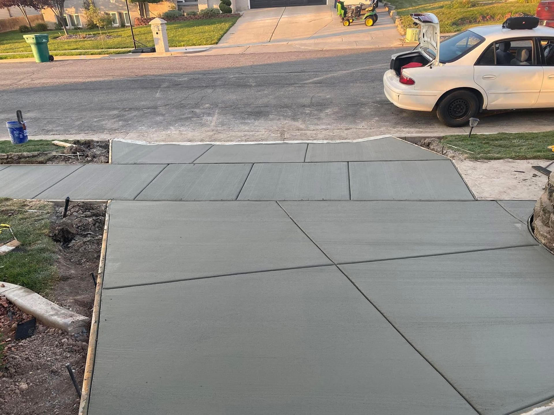 Freshly poured gray concrete driveway and sidewalk sections outside a house with a white sedan parked at the curb.
