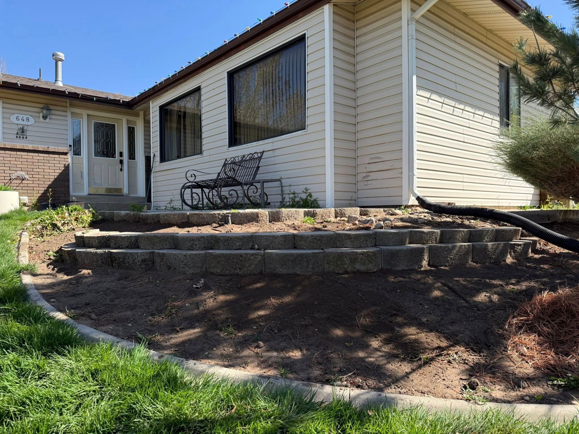 A beige house with a curved, multi-tiered stone retaining wall and a small iron bench on a stone patio.