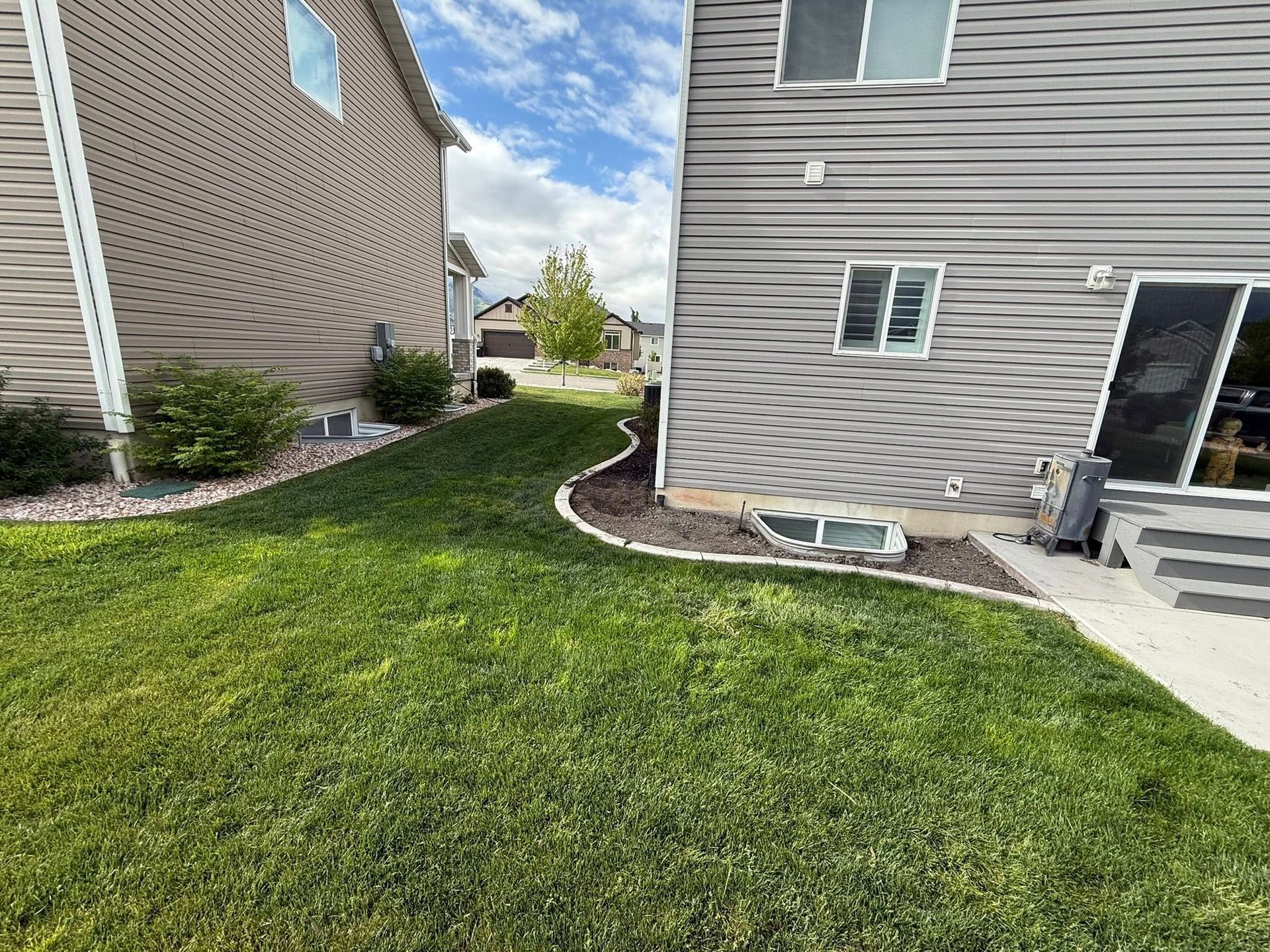 The side yard of a suburban house, featuring a grassy lawn, a concrete pathway, and landscaping along the siding.