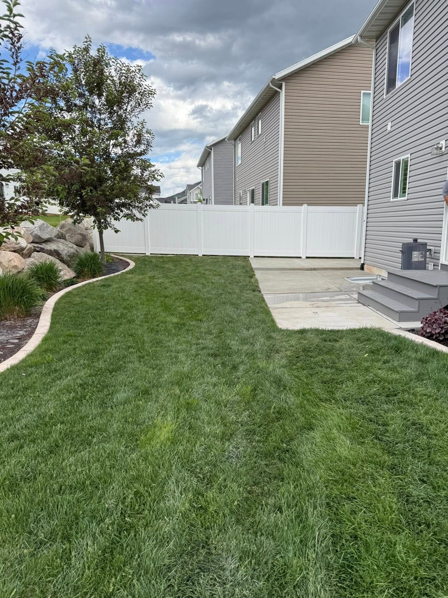 A backyard with a vibrant green lawn, a white vinyl fence, a stone landscaping border, and a concrete patio by a house.