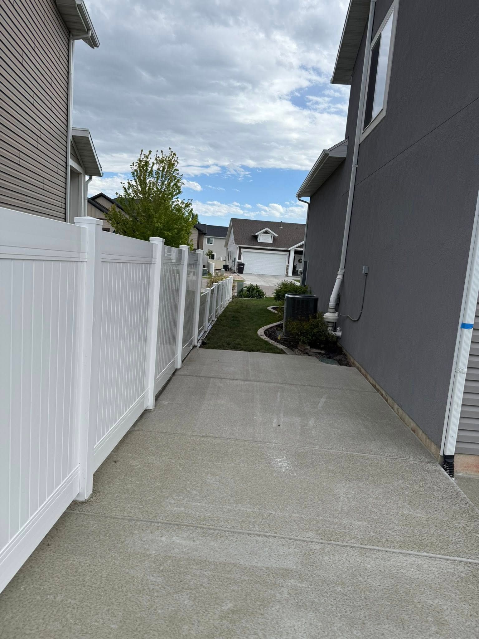 A narrow concrete path between a tall white vinyl fence and the side of a grey house, leading toward other homes.