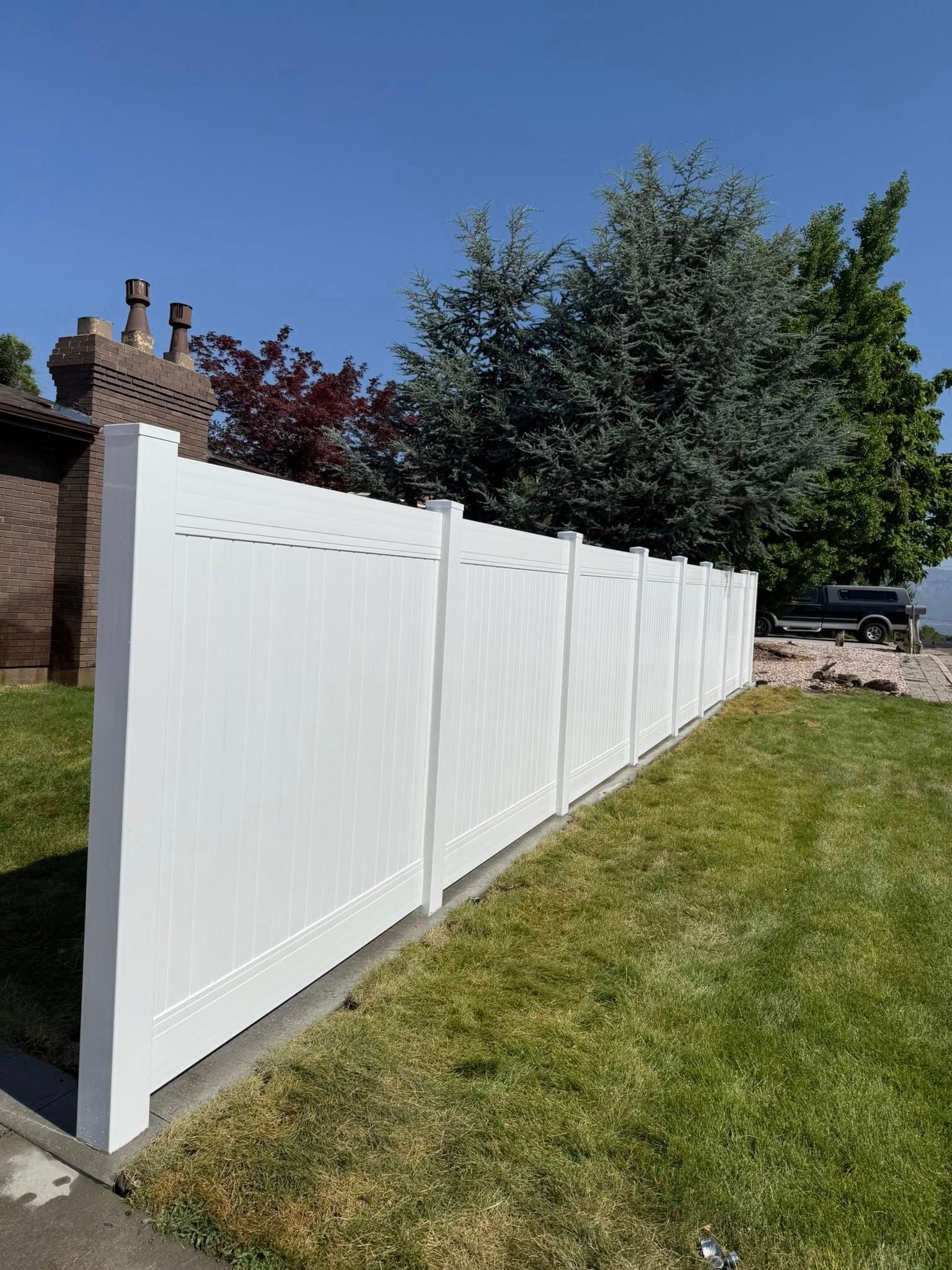 A white vinyl privacy fence stands on a mowed lawn next to a brick house and green trees under a clear blue sky.
