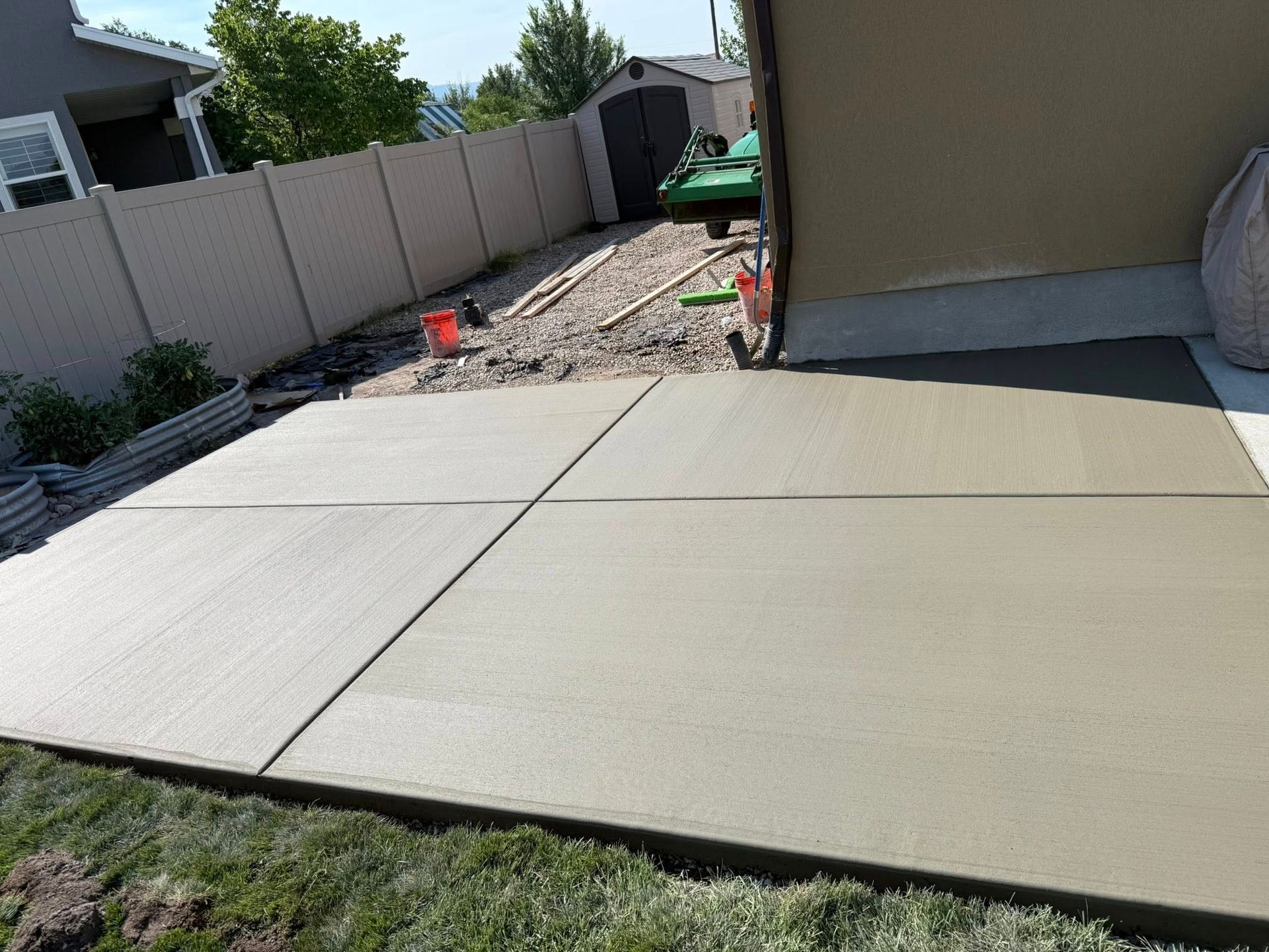 A freshly poured rectangular concrete patio in a backyard next to a house with a tan fence and a small shed in the back.