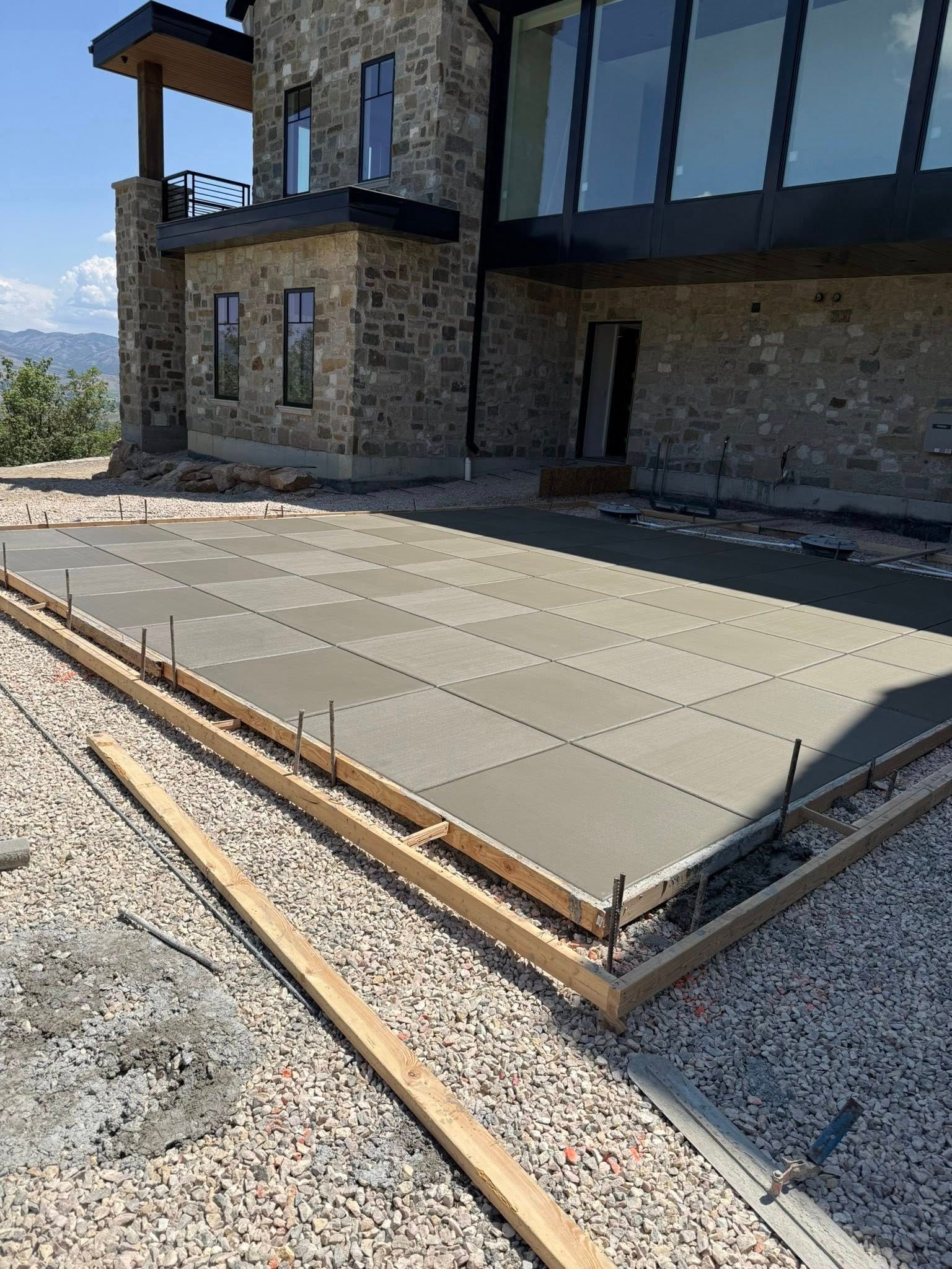 A freshly poured concrete patio section with a grid pattern, framed by wooden forms in front of a modern stone house.