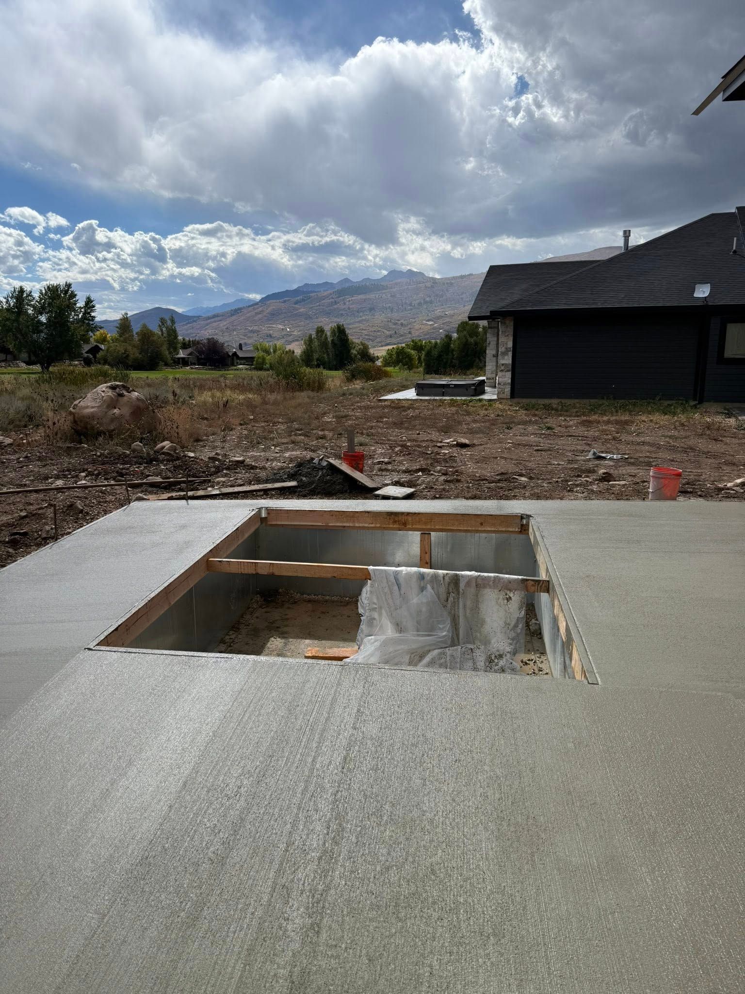 A rectangular cutout in a newly poured concrete patio slab with wooden framing visible beneath in a mountain setting.
