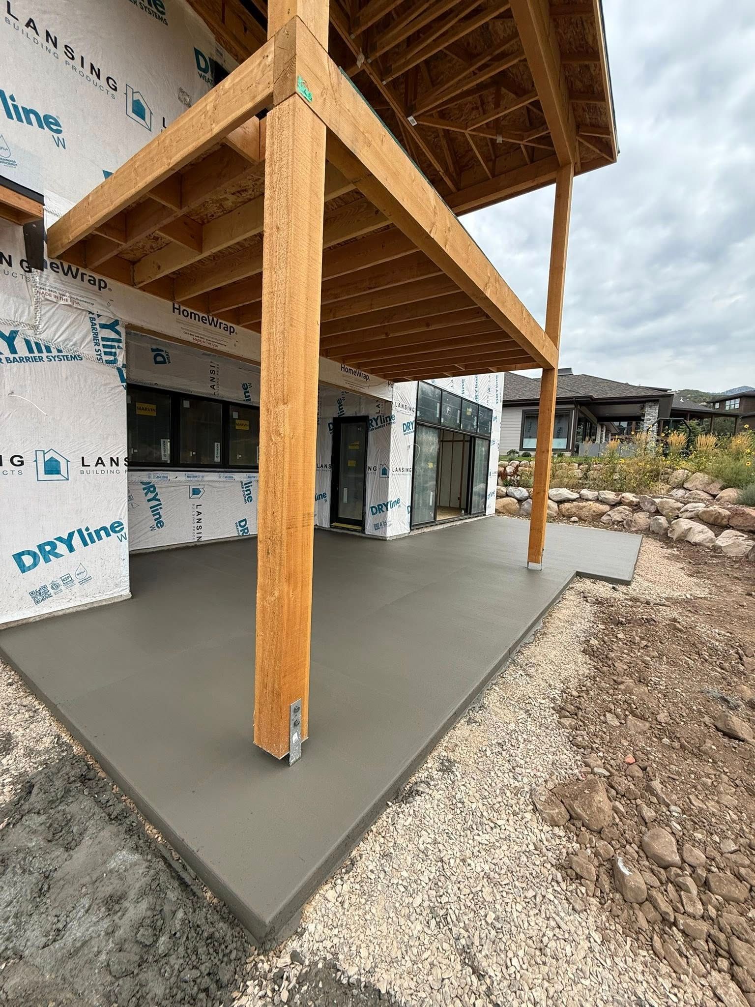 A freshly poured concrete patio under a wooden deck attached to a house currently under construction.