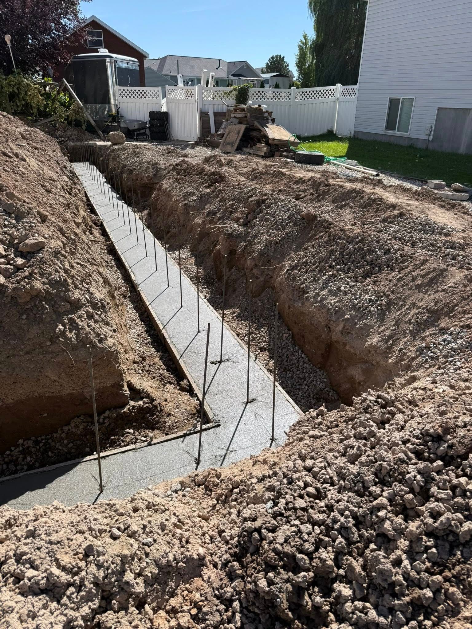 A freshly poured concrete footing for a foundation, centered in a deep excavation trench under a bright, sunny sky.